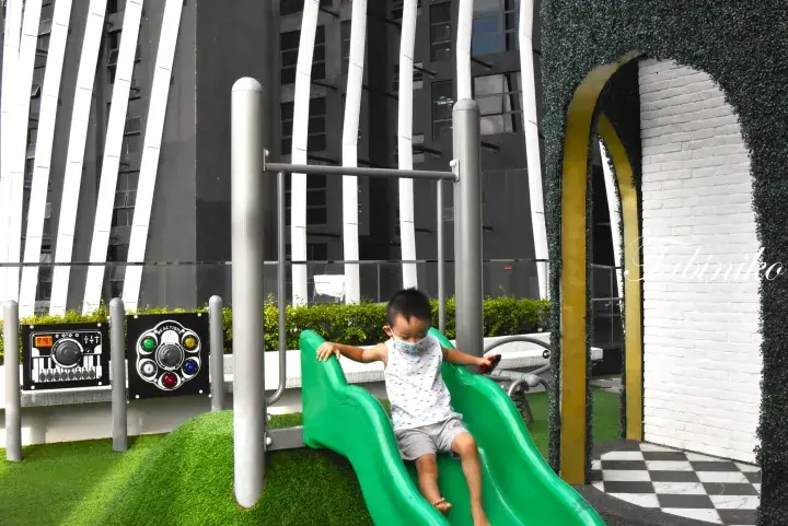 A toddler slides down a green slide at a modern playground with musical panels and a checkered floor.
