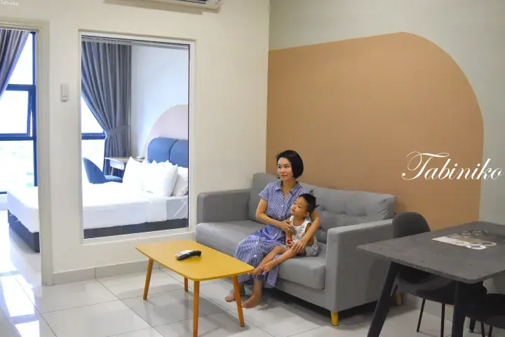 A woman and child relax on a sofa in a modern serviced apartment living room with a view of the bedroom.