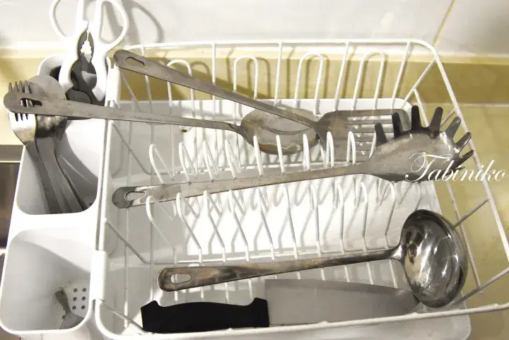 Assorted stainless steel kitchen utensils, including pasta servers and a ladle, drying in a white dish rack.