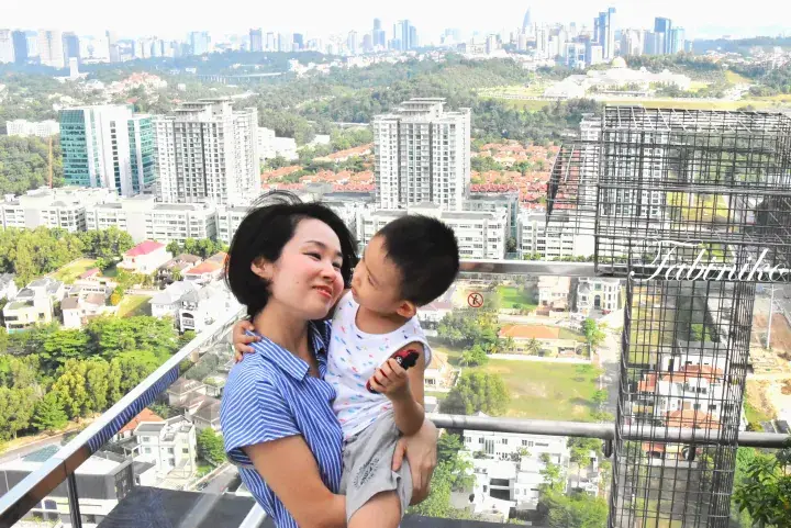 Mother and child on a high-rise balcony overlooking lush greenery and the Kuala Lumpur skyline.