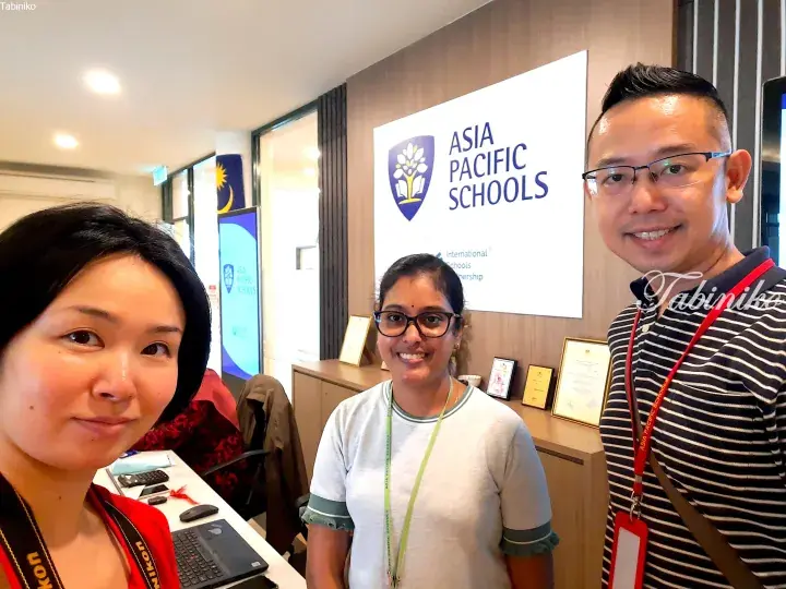 Three people smiling in front of the Asia Pacific Schools sign at an education event in Malaysia.