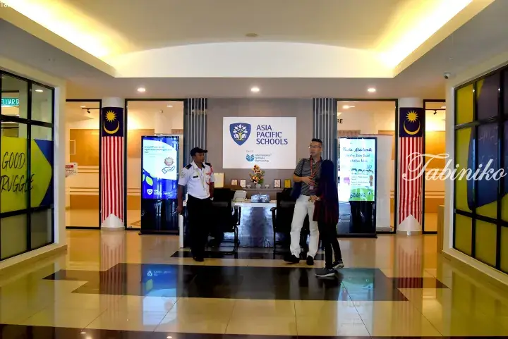 Entrance to Asia Pacific Schools Malaysia with security guard and visitors near Malaysian flags.
