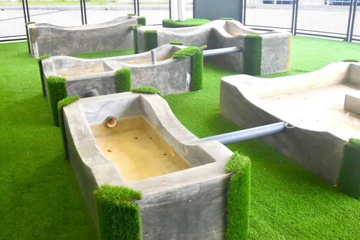 Concrete water play structures with artificial grass in an outdoor children&rsquo;s playground area.
