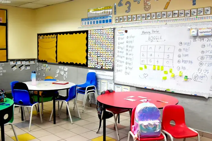 Bright classroom with colorful tables and chairs, featuring a whiteboard with a word game and educational posters on the walls.