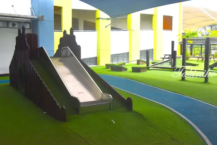 Children&rsquo;s playground featuring a stainless steel slide and climbing structure on artificial grass with a blue running track.