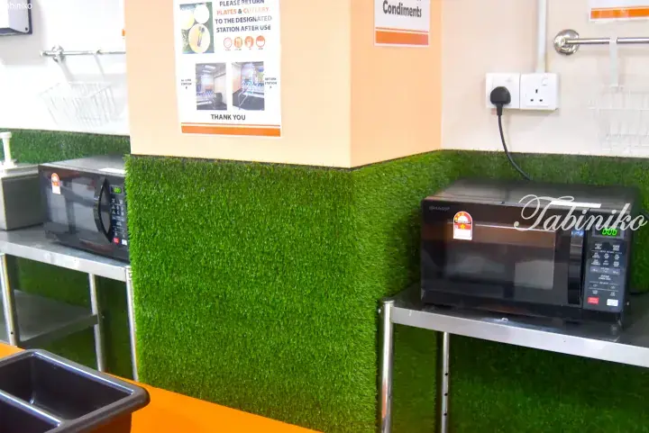 Microwave ovens on stainless steel shelves with green artificial grass wall in a Malaysian food court.