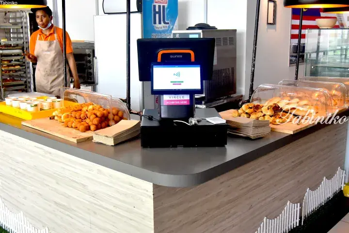 A bakery counter displays golden fried snacks and pastries behind a digital payment terminal, with a staff member in an orange shirt and apron visible in the background.
