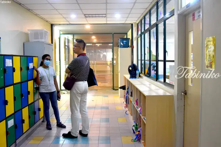 Man and woman standing in a school hallway with colorful lockers and a low wooden shelf near a window.