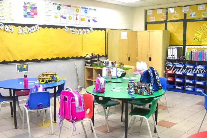 Colorful classroom with round tables, chairs, and children&rsquo;s backpacks, featuring yellow bulletin boards and educational displays.