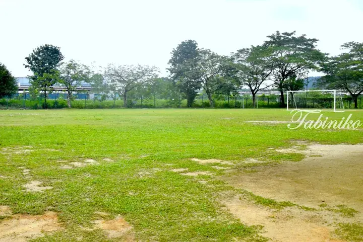 Grassy football field with goalposts and trees in the background, likely a local park or school ground in Malaysia.