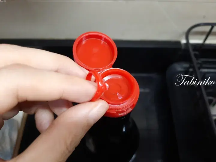 Close-up of an open red plastic cap on a dark glass bottle, likely containing soy sauce or vinegar, on a black countertop.
