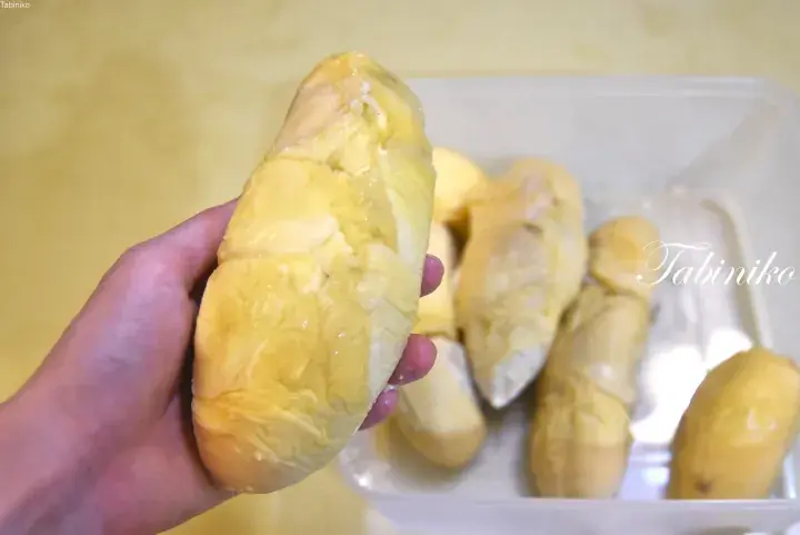 A hand holding a large piece of fresh durian flesh with more pieces in a container.