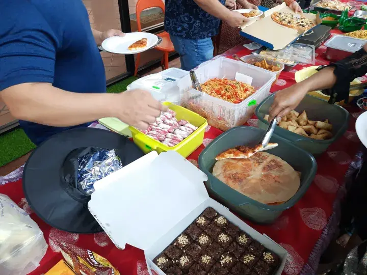 Guests serving themselves from a buffet table featuring pizza, samosas, brownies, and pasta at a Malaysian gathering.
