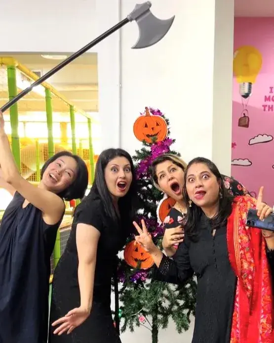 Four women pose playfully with a prop axe near a Halloween-decorated tree in a colorful indoor play area.