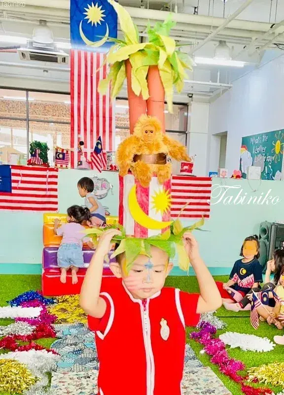 Child in red outfit with face paint holds a handmade palm tree prop at a Malaysian Independence Day school celebration.
