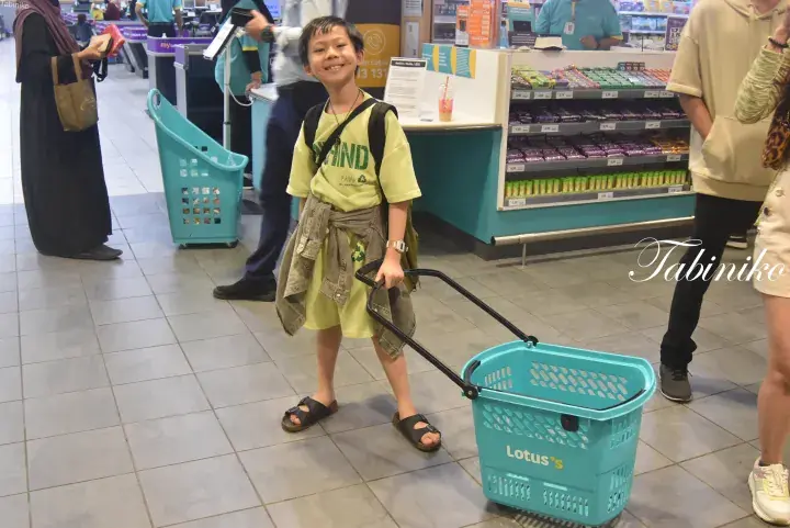 Young boy smiling while pulling a blue Lotus’s shopping basket in a Malaysian supermarket.