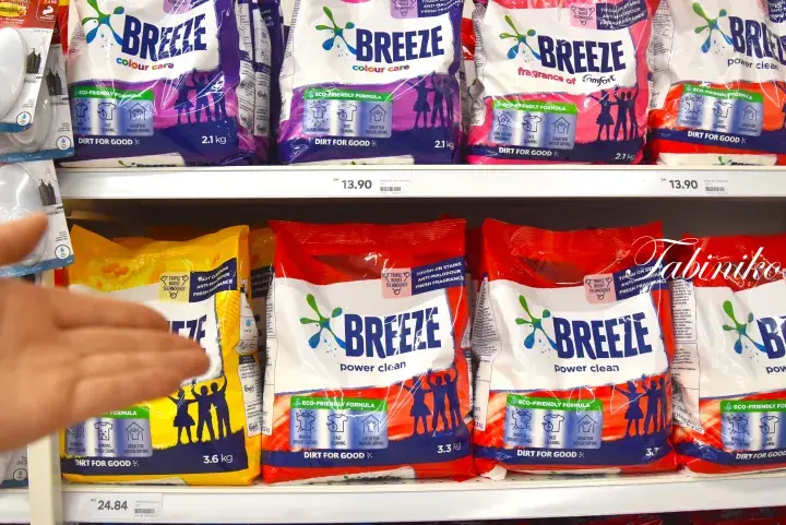 A hand reaches for a yellow bag of Breeze laundry detergent on a supermarket shelf alongside other Breeze product variants.
