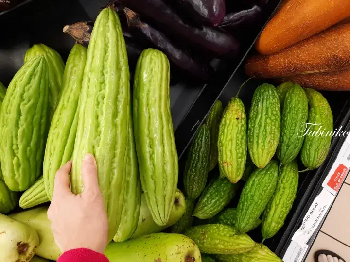 Hand selecting long bitter melons in a Malaysian supermarket vegetable section alongside eggplants and carrots.