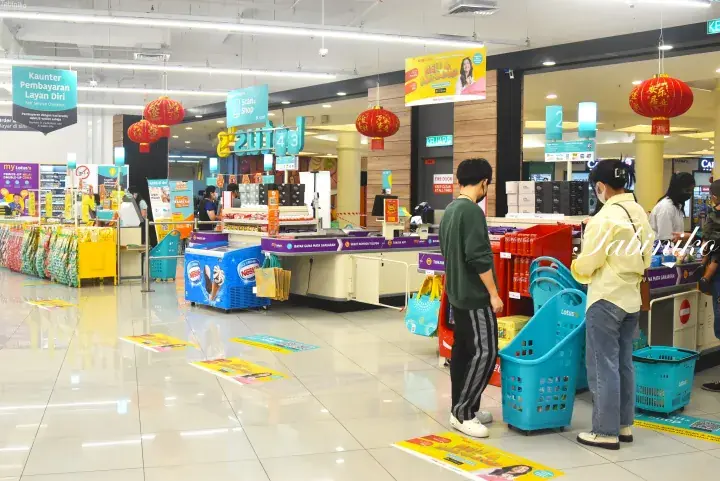 Shoppers with blue baskets queue at a supermarket checkout in Malaysia, decorated with festive red lanterns.