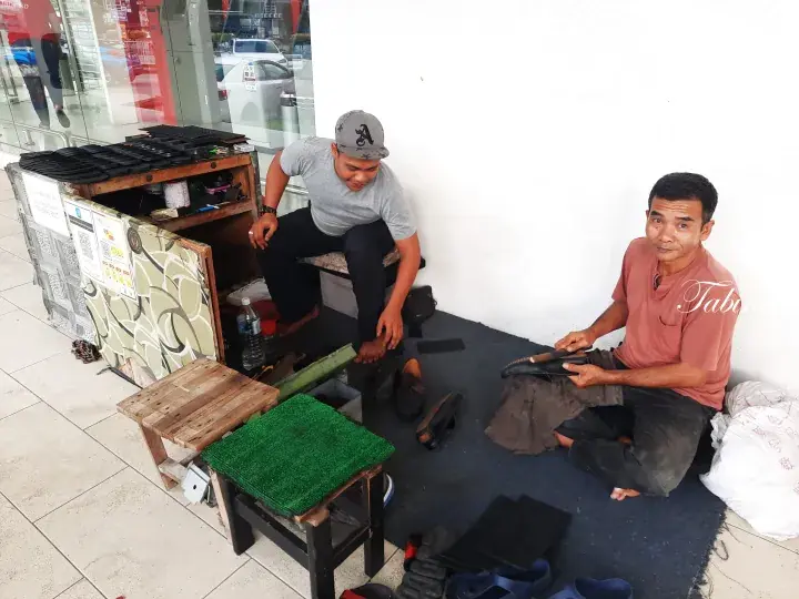 Two shoe shiners at a roadside stall in Malaysia, one cleaning a shoe while the other waits for customers.