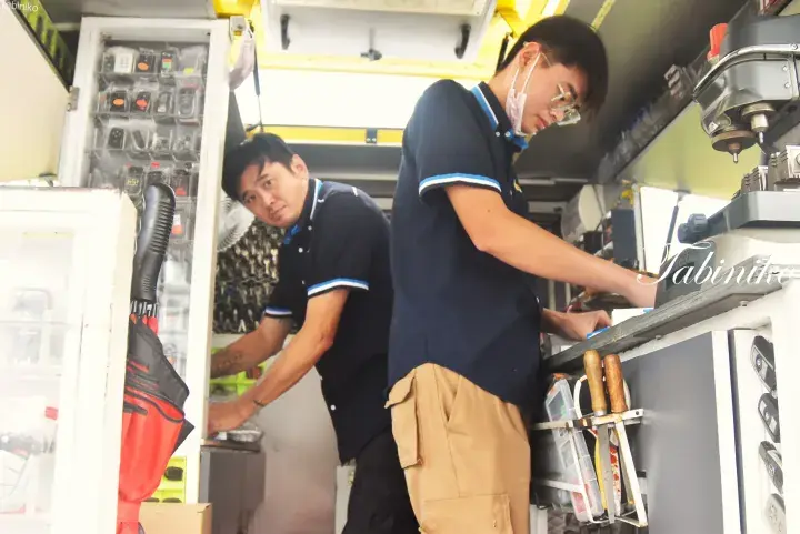 Two technicians in uniform working inside a mobile workshop van, one using a drill press while the other organizes tools.