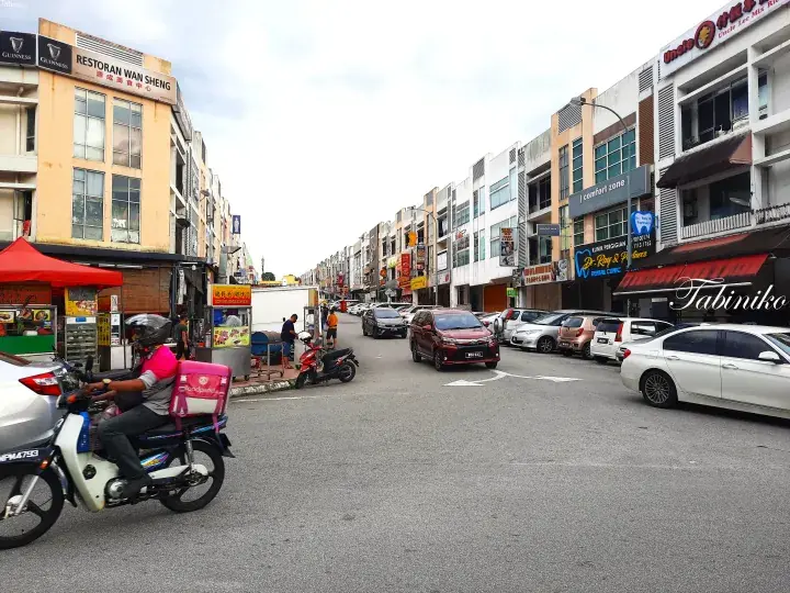 A street view of a Malaysian commercial area with a food delivery rider, parked cars, and shophouses.
