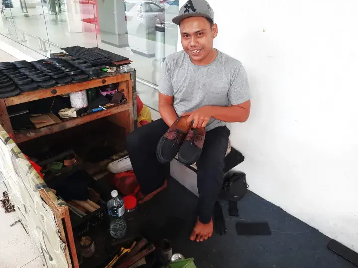 A shoe shiner sits with his tools and a pair of shoes at a street stall in a Malaysian shopping mall.