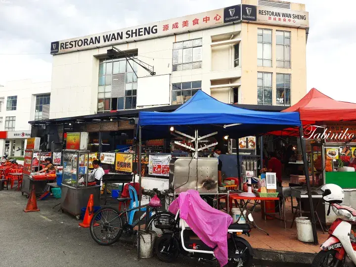 Street food stalls under blue and red tents outside Restoran Wan Sheng food court in Malaysia.