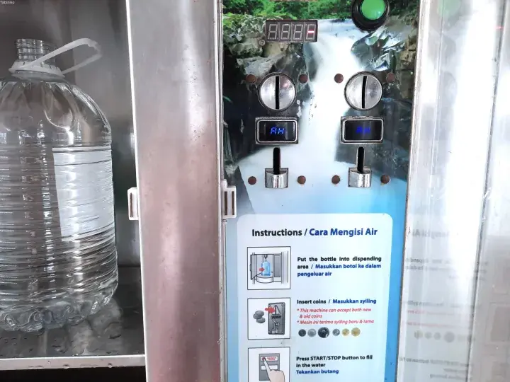 A large water bottle being filled at a coin-operated water dispenser with bilingual instructions.