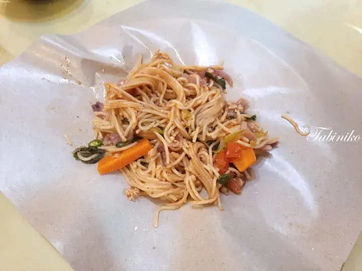 A serving of stir-fried noodles with vegetables and meat on a paper wrapper, typical of Malaysian hawker centre fare.