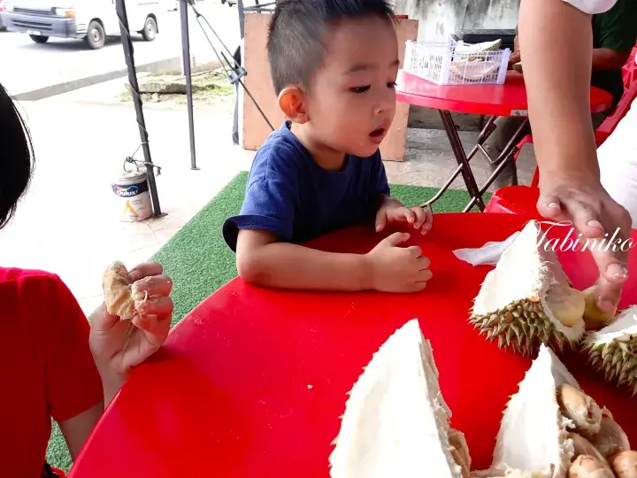 A young boy sits at a red table enjoying durian fruit at a Malaysian roadside stall.