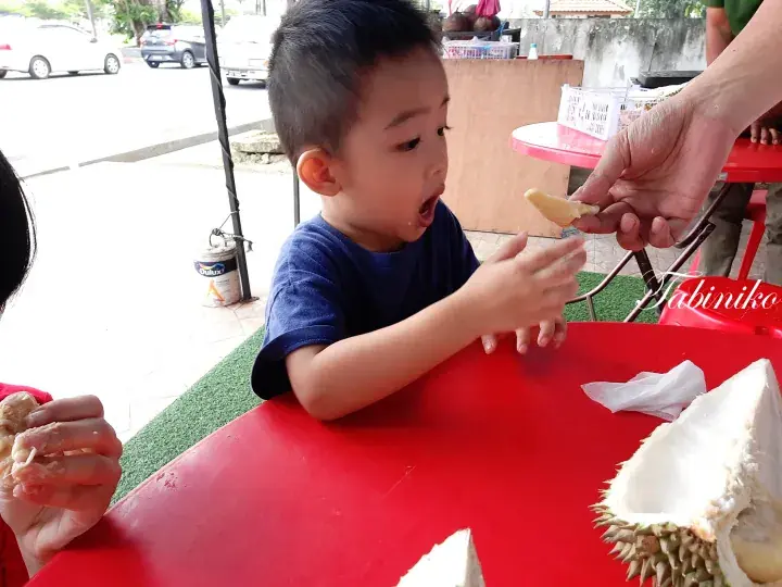 A young boy eagerly reaching for a piece of durian at a red table with a durian shell nearby.