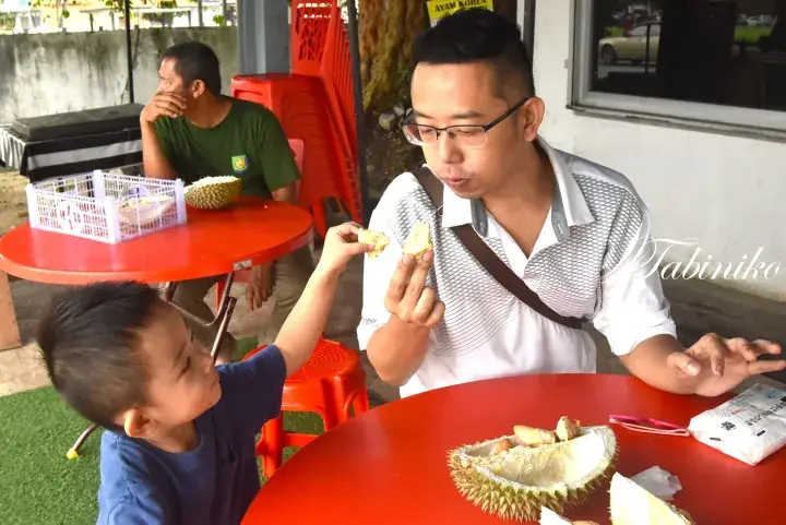A young boy offers durian to a man at a red table, with another man eating durian in the background.