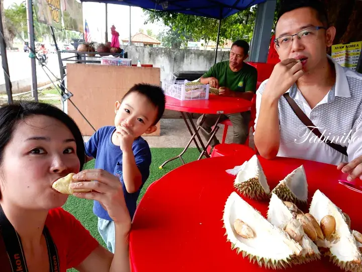 A family enjoys fresh durian at a red table under a blue tent at a Malaysian roadside stall.