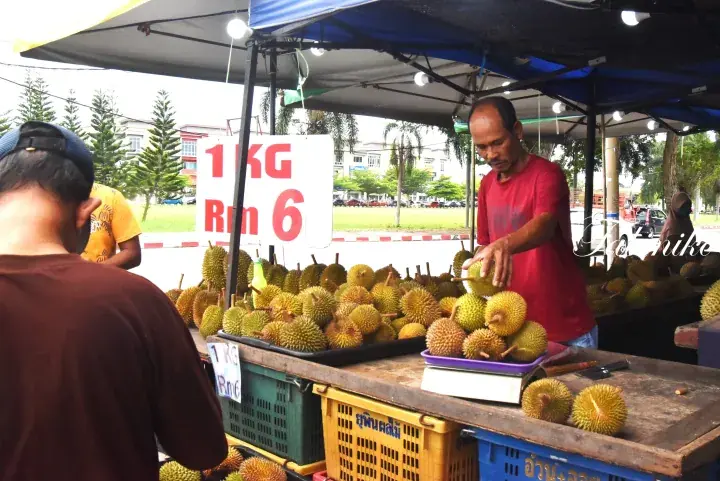 A vendor sells durian at an outdoor market stall with a sign showing 1KG for RM 6 in Malaysia.