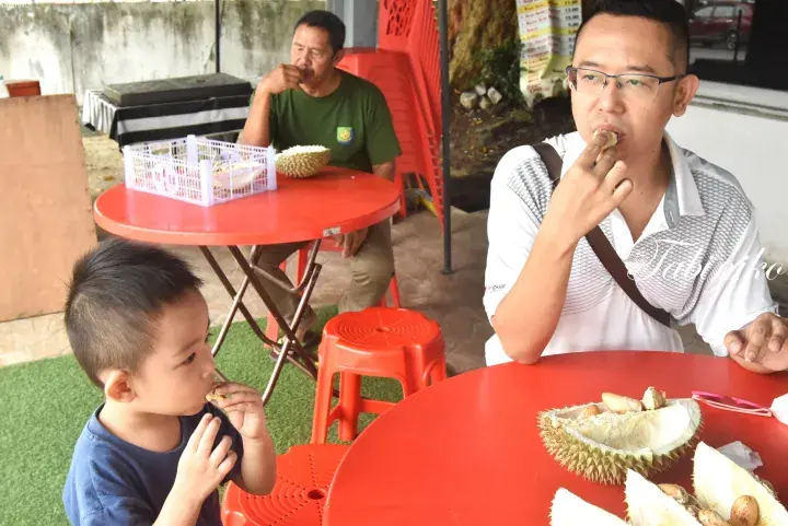 A family enjoys durian at a roadside stall in Malaysia, with a child and two men eating the fruit at red tables.