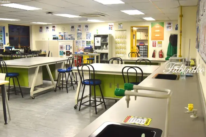 Modern science laboratory with green-topped workstations, blue stools, and safety equipment in a well-lit room.