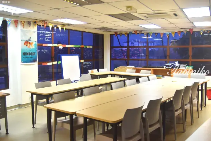 Modern co-working space with long wooden tables, grey chairs, and large windows overlooking a city view.