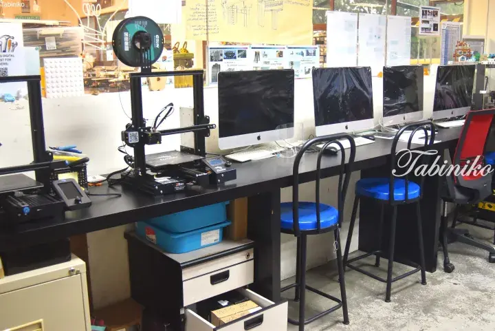 A workspace featuring a 3D printer and multiple Apple computers on a long table with blue stools.