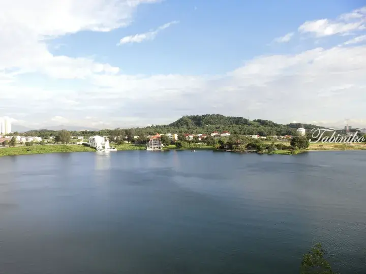 Scenic view of a lake in Malaysia with waterfront homes and a green hill under a blue sky.