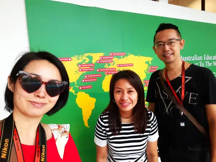 Three people pose in front of a green wall featuring a yellow world map and Australian education information.