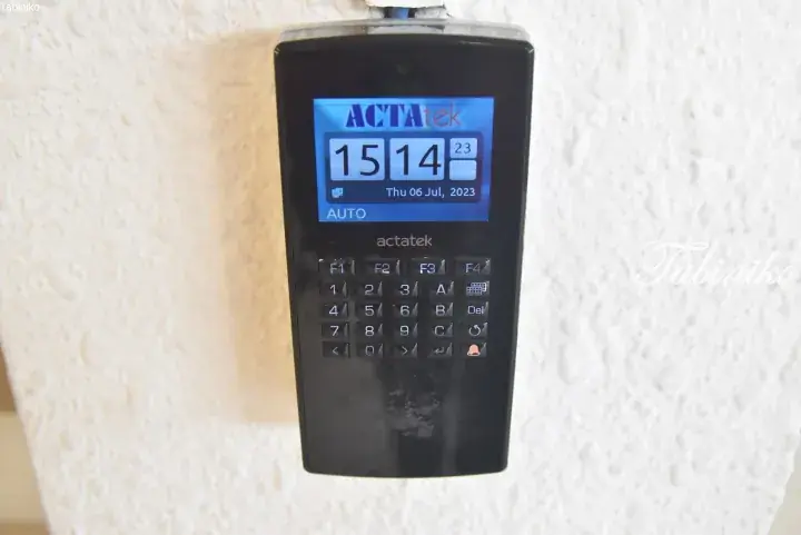 Black ACTAtek digital time attendance clock mounted on a textured white wall showing the date July 6, 2023.