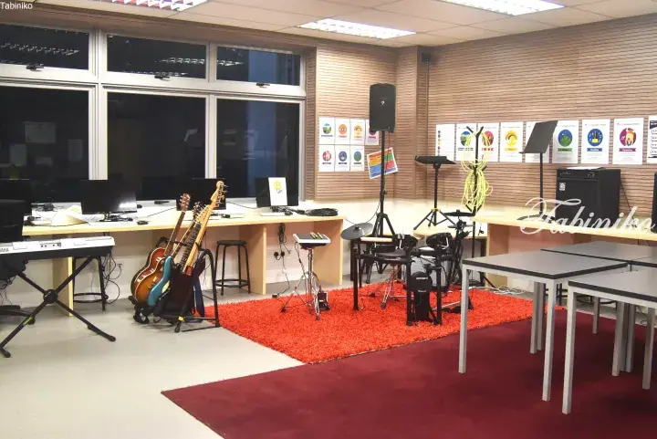 Music practice room with guitars, keyboard, drum kit, and computers in a bright studio space.