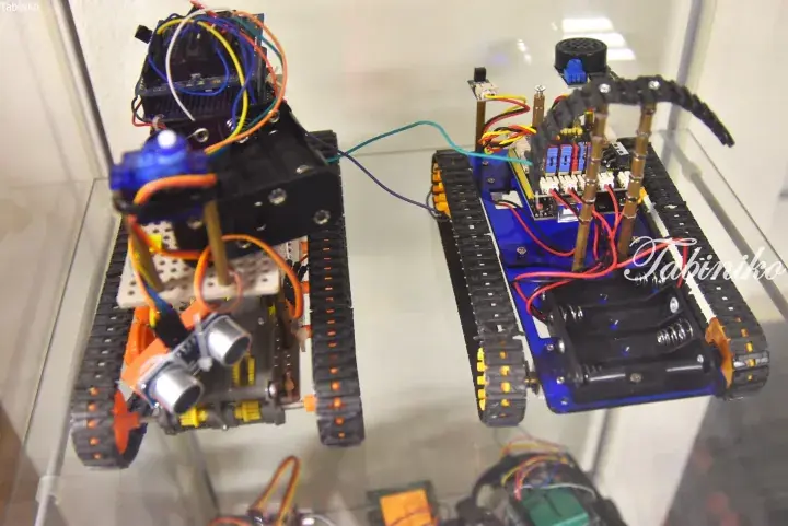 Two DIY tracked robots with exposed circuitry and sensors displayed on a glass shelf.