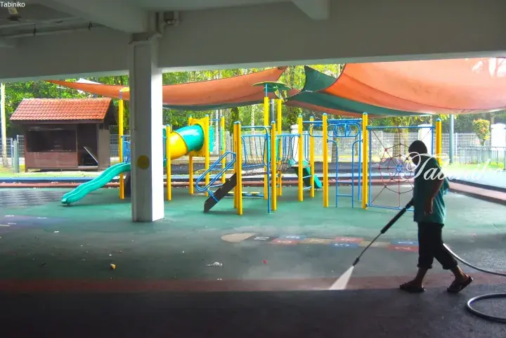 A worker pressure-washing the rubber flooring of a colorful children&rsquo;s playground under a shaded structure.