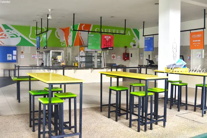 Bright cafeteria with yellow tables and green stools, featuring food service counters and menu boards.