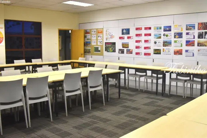 Modern meeting room with U-shaped tables, white chairs, and a wall display of colorful brochures and posters.