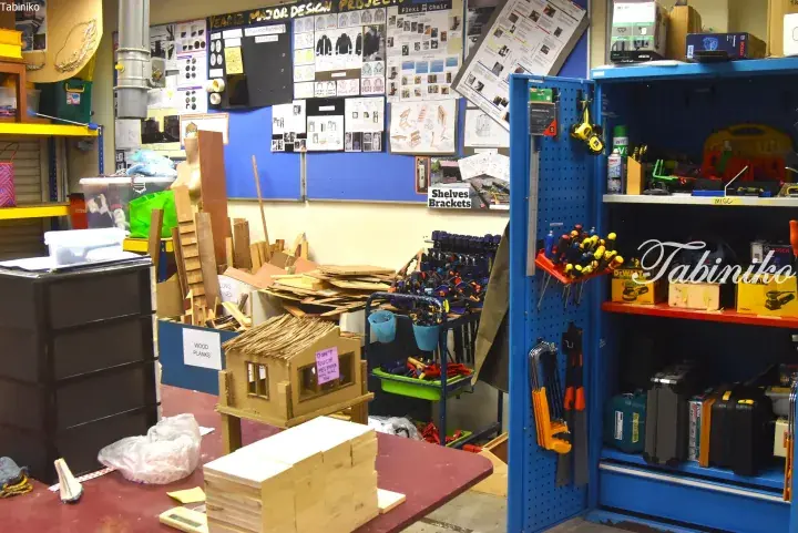 A well-equipped workshop with blue shelving, tools, wood pieces, and design boards on the wall.