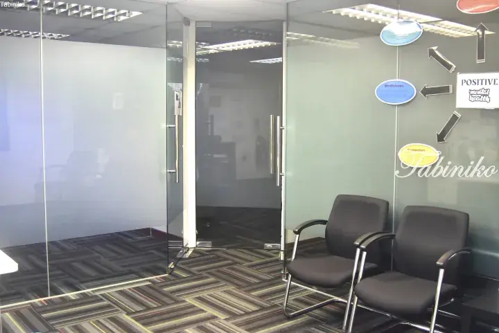 Modern office interior with glass partition walls, frosted panels, and two black chairs in the waiting area.