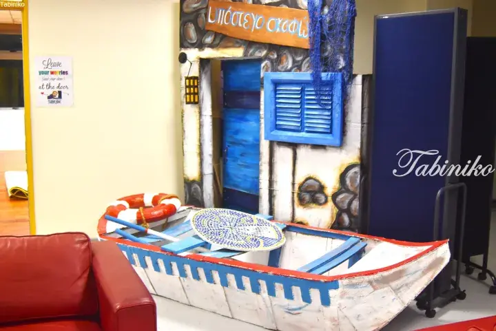A decorative boat prop inside a Greek-themed room with a painted cottage facade and blue shutters.
